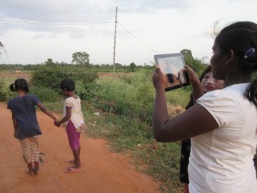 Image showing two children holding hands and one person with an iPad filming. One further person looks on.